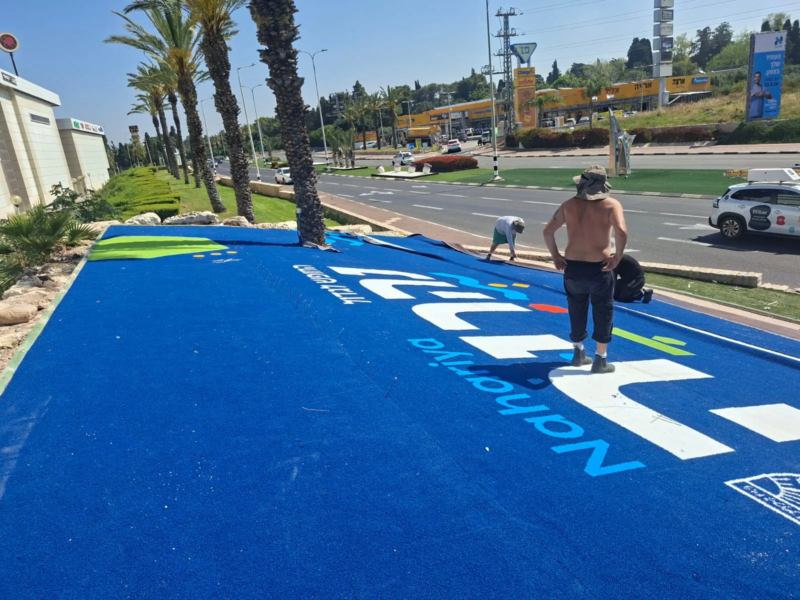 ColorPlay installer positioning the large blue Nahariya turf sign on a roadside traffic island lined with palm trees