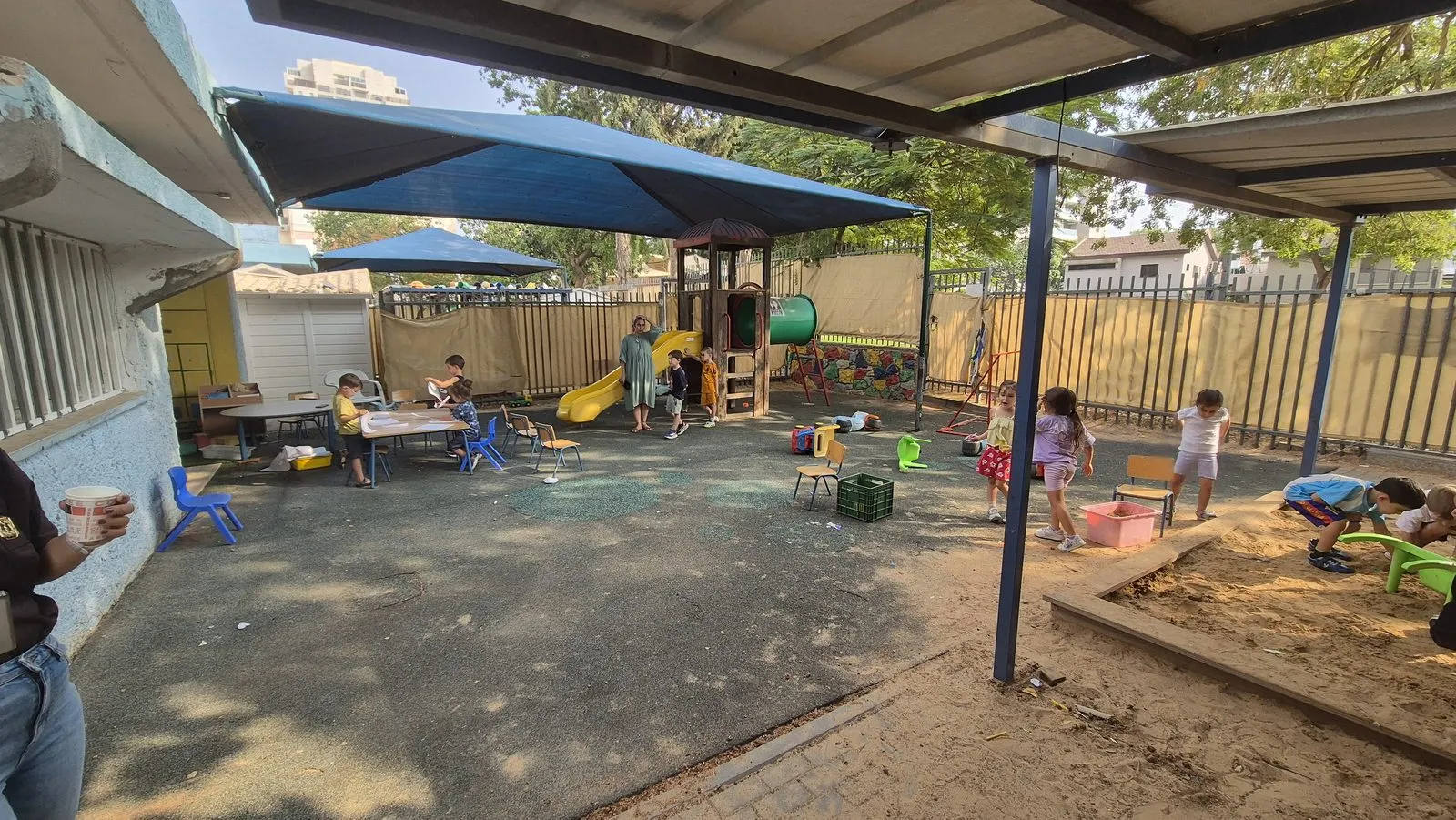 The kindergarten yard before the work: a worn dark rubber surface with a play structure, shade canopies and scattered chairs