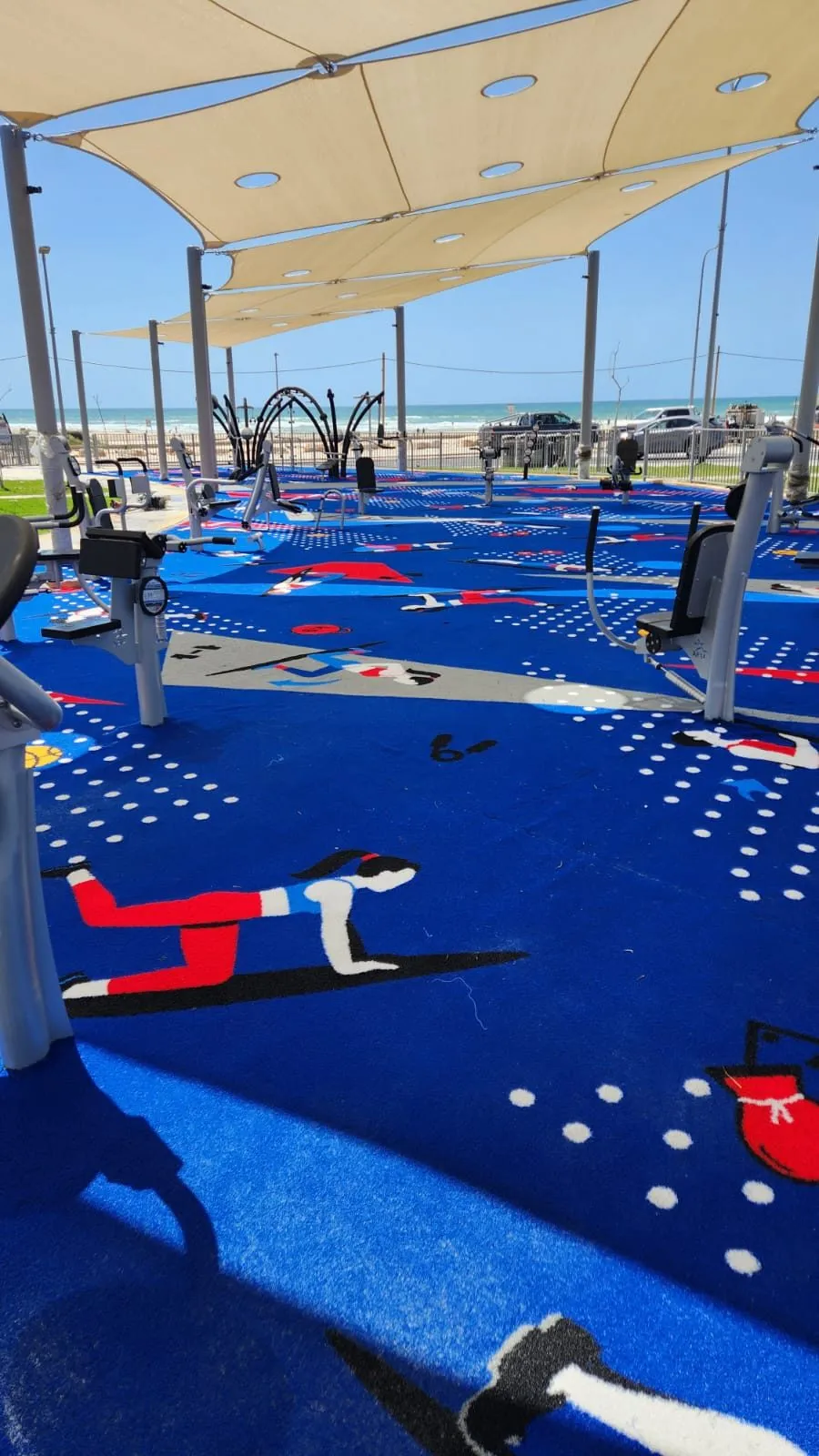 Outdoor gym machines set on the blue ColorPlay surface with exercise figures and the beach and sea in the background