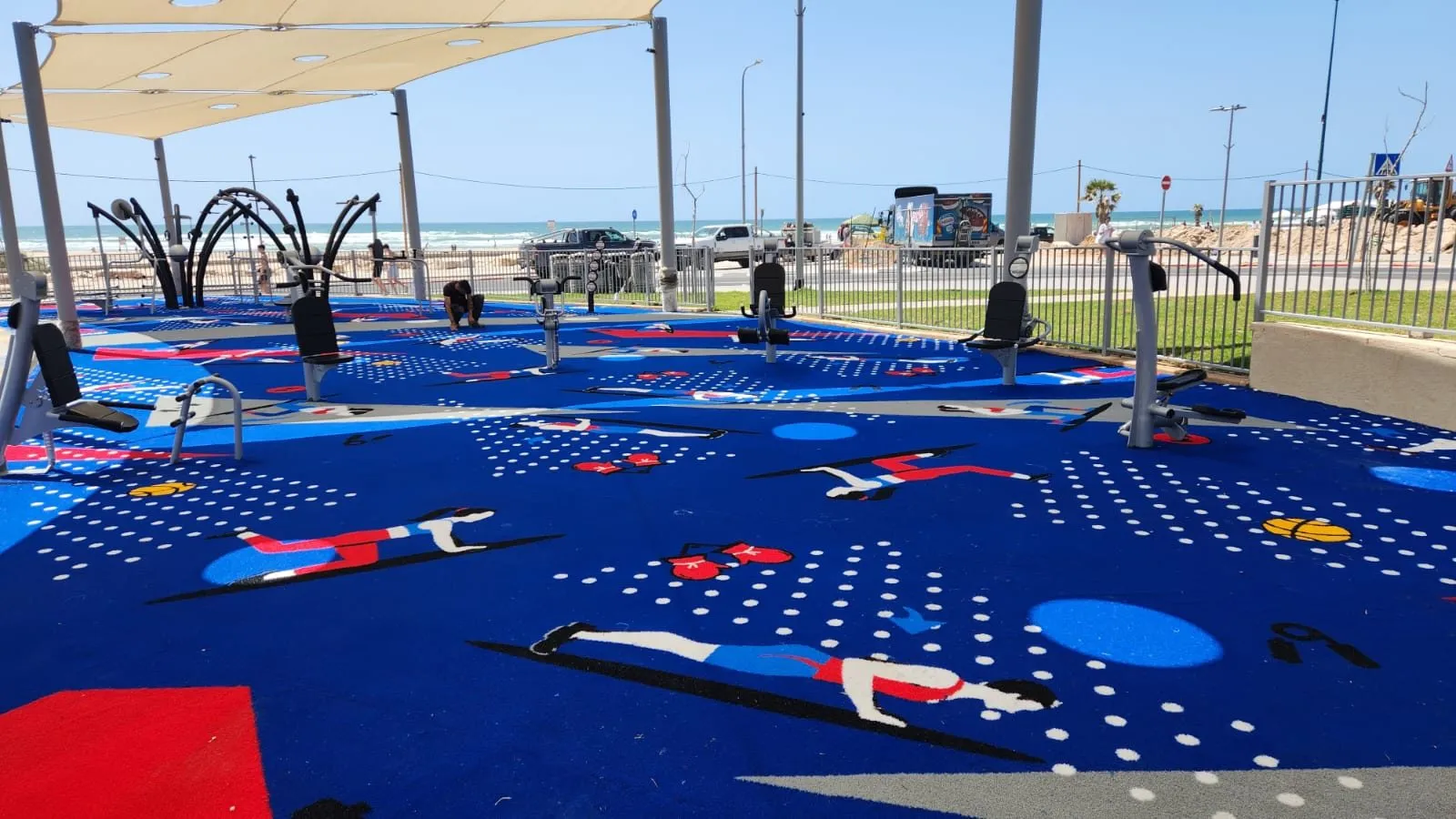 Blue ColorPlay outdoor-fitness surface with exercise figures, gym machines and shade sails beside the sea at Poleg Beach