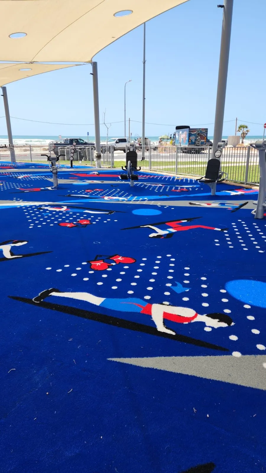 Plank and exercise figures on the blue surface beneath beige shade sails, with the promenade and sea beyond