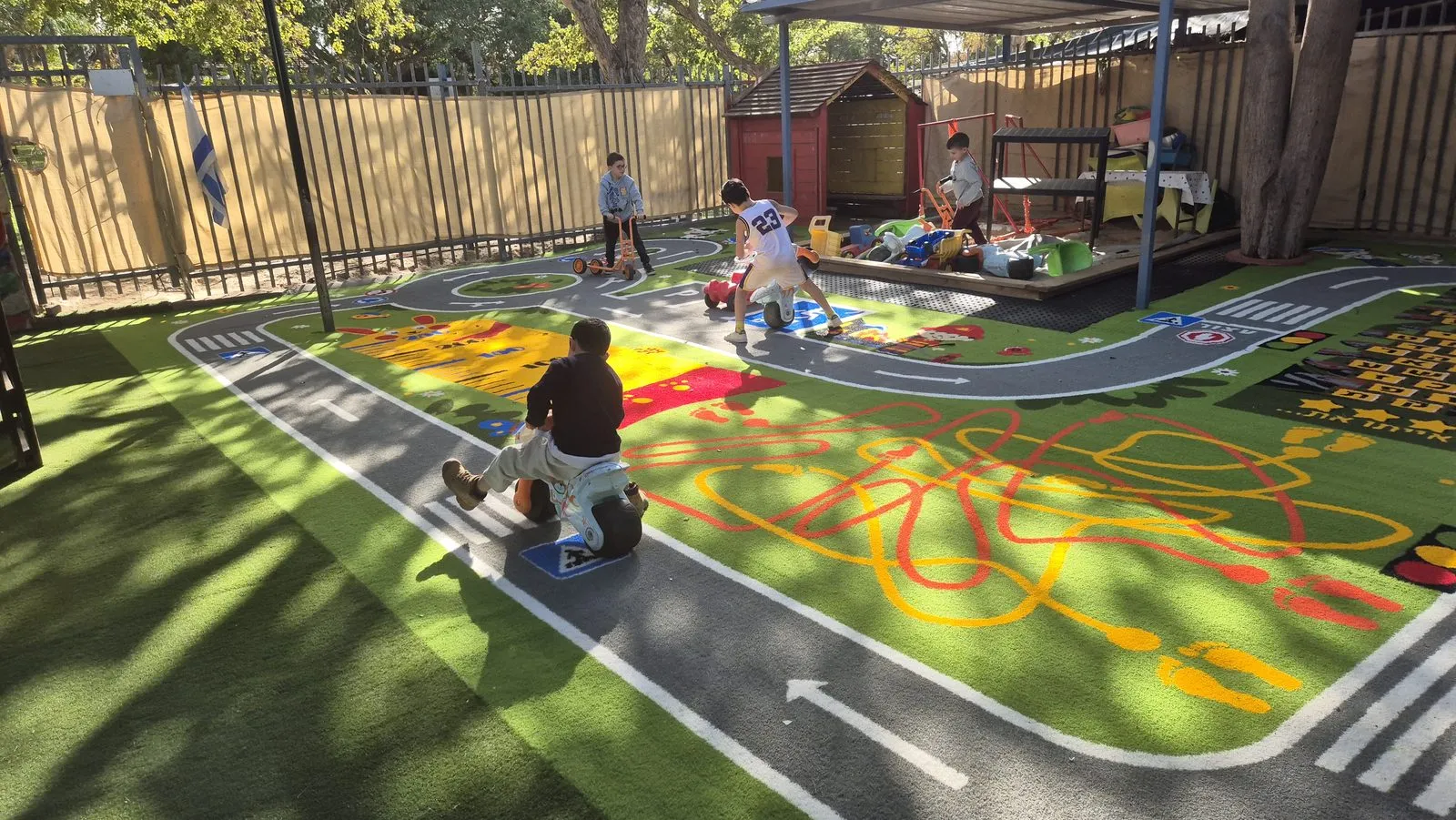 Kindergarten yard resurfaced in green artificial grass with a road circuit, hopscotch and squiggle trails, with children riding scooters