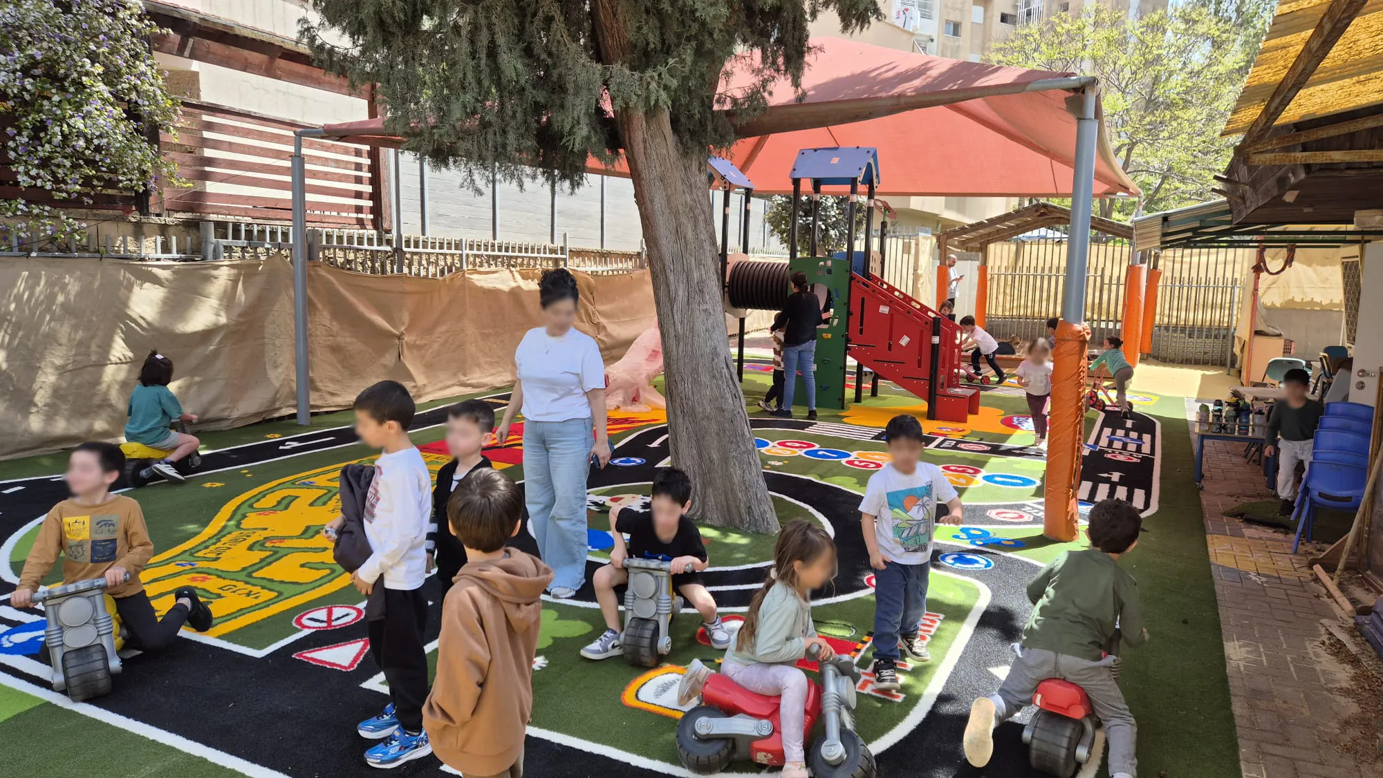 Children riding toy cars around the road circuit on the ColorPlay turf surface beside the tree